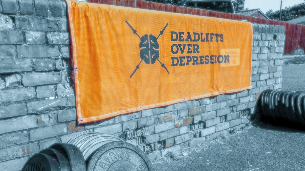 The Orange Deadlifts Over Depression Banner sitting proudly amongst weight plates at Deadlifts Over Depression, Cambridgeshire