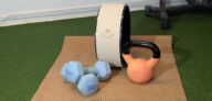 Myga Yoga setup with sustainable cork yoga mat, cast iron kettlebell, yoga wheel, and light blue dumbbells on gym turf.