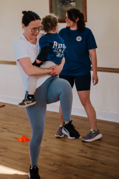 Woman holding a young child while completing a standing knee raise during a FitFactory fitness class in Sawtry.